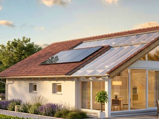 Modernes Bausatzhaus von Bio-Solar-Haus mit Photovoltaik-Modulen und großflächiger Verglasung zur goldenen Stunde mit Terrasse mit Loungemöbeln, umgeben von einer gepflegten Gartenanlage und Bäumen unter einem leicht bewölkten Himmel.
