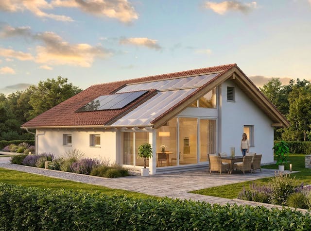 Modernes Bausatzhaus von Bio-Solar-Haus mit Photovoltaik-Modulen und großflächiger Verglasung zur goldenen Stunde mit Terrasse mit Loungemöbeln, umgeben von einer gepflegten Gartenanlage und Bäumen unter einem leicht bewölkten Himmel.