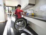 Barrierefrei bauen, Frau im Rollstuhl in einer großzügigen und offenen Küche, im Hintergrund trägt ein Mädchen ein Tablett, Foto: KfW-Bildarchiv / photothek