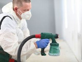 Spritzpistole, Bild eines Mannes im Schutzanzug und mit Atemmaske und einer professionellen Farb-Spritzpistole, mit der er die Wand weiß ansprüht, Foto: H_Ko / stock.adobe.com