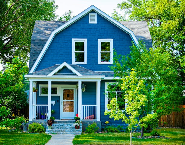 Blaues amerikanisches Holzhaus mit Mansarddach, das mit den kennzeichnenden Holzschindeln gedeckt ist. Eine gemütliche Veranda, Schiebefenster und Rahmentür runden das Bild stilecht ab.