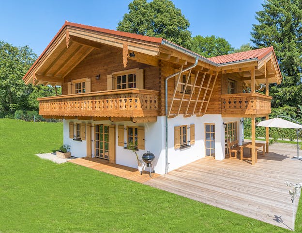 Modernes Chalet in Holzmassivbauweise mit weitem Dachüberstand, verziertem Holzbalkon und heller Putzfassade im Erdgeschoss von Rems-Murr-Holzhaus. Besonders ins Auge fallen liebevolle Details wie die Eckverbindungen als sogenanntes Tiroler Schloss oder das Schnitzwerk an den Balkonbrüstungen.